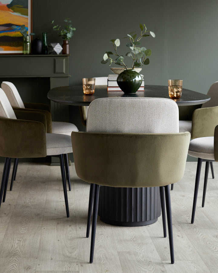 A round dining table with a dark fluted base is surrounded by modern chairs with olive green and cream upholstery.