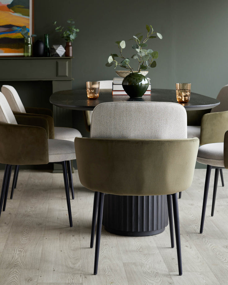 A round dining table with a dark fluted base is surrounded by modern chairs with olive green and cream upholstery.