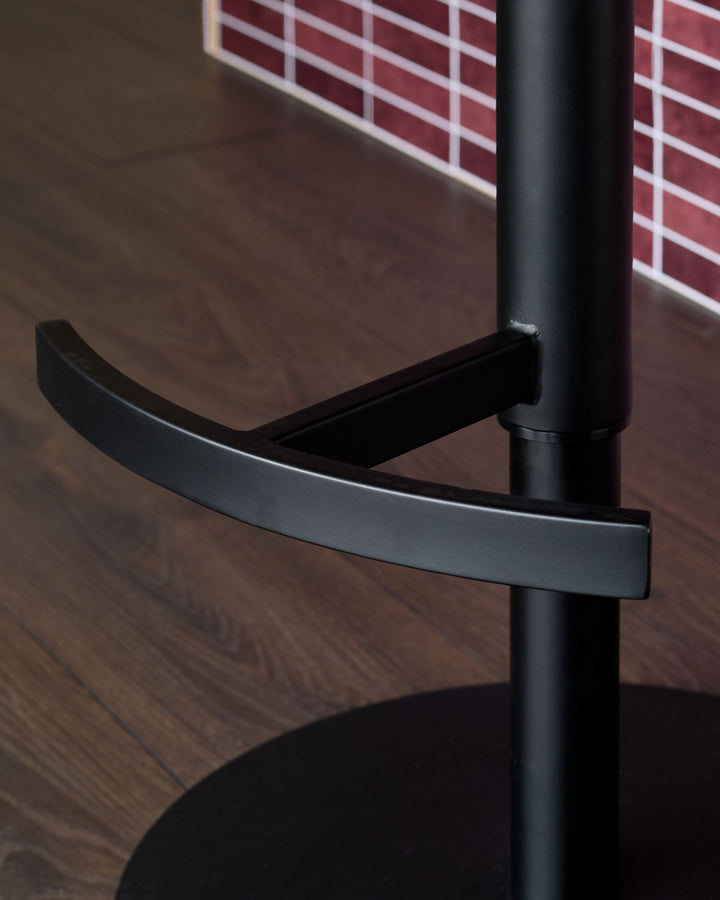 Black gas-lift bar stool with curved footrest and cylindrical pole base, shown in close-up detail.