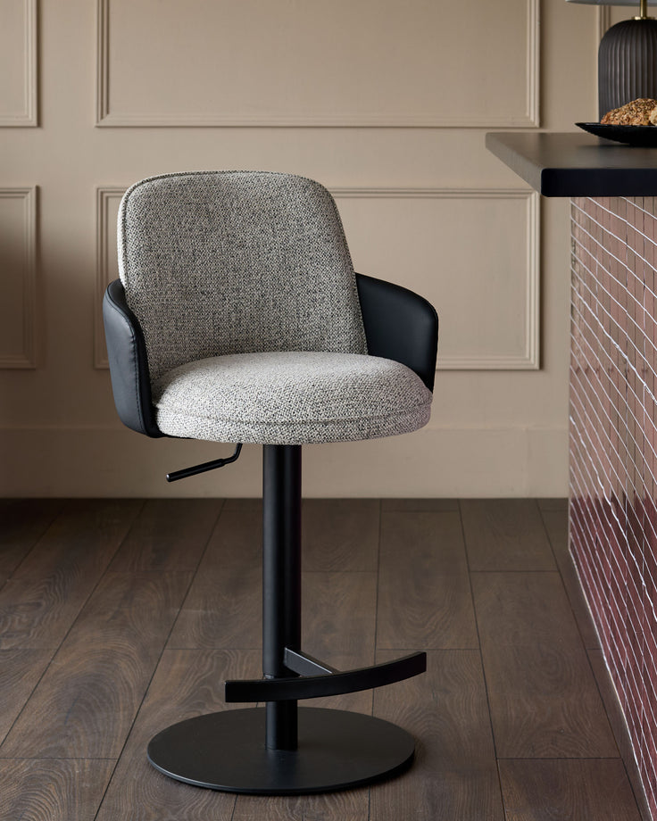 Wren gas lift bar stool in grey fabric with black faux leather armrests and black metal base, shown in neutral interior setting.