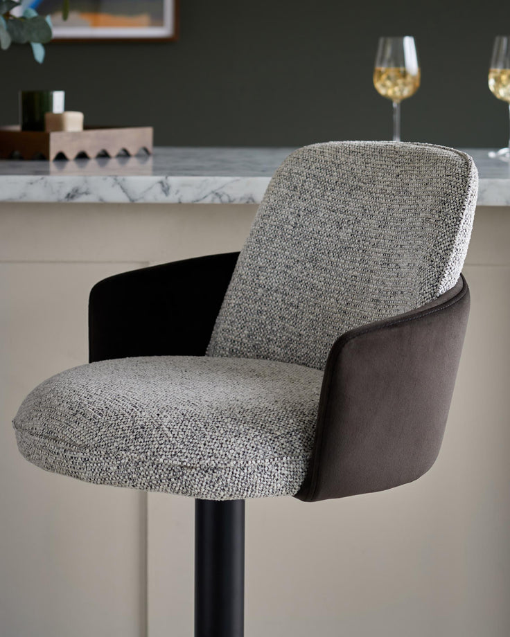 Grey tweed bar stool with dark brown arms and a black stand, placed against a bar setting with wine glasses.