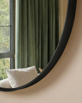 Black curved wall mirror with organic, flowing shape displayed above a cream upholstered chair near a window with green curtains.