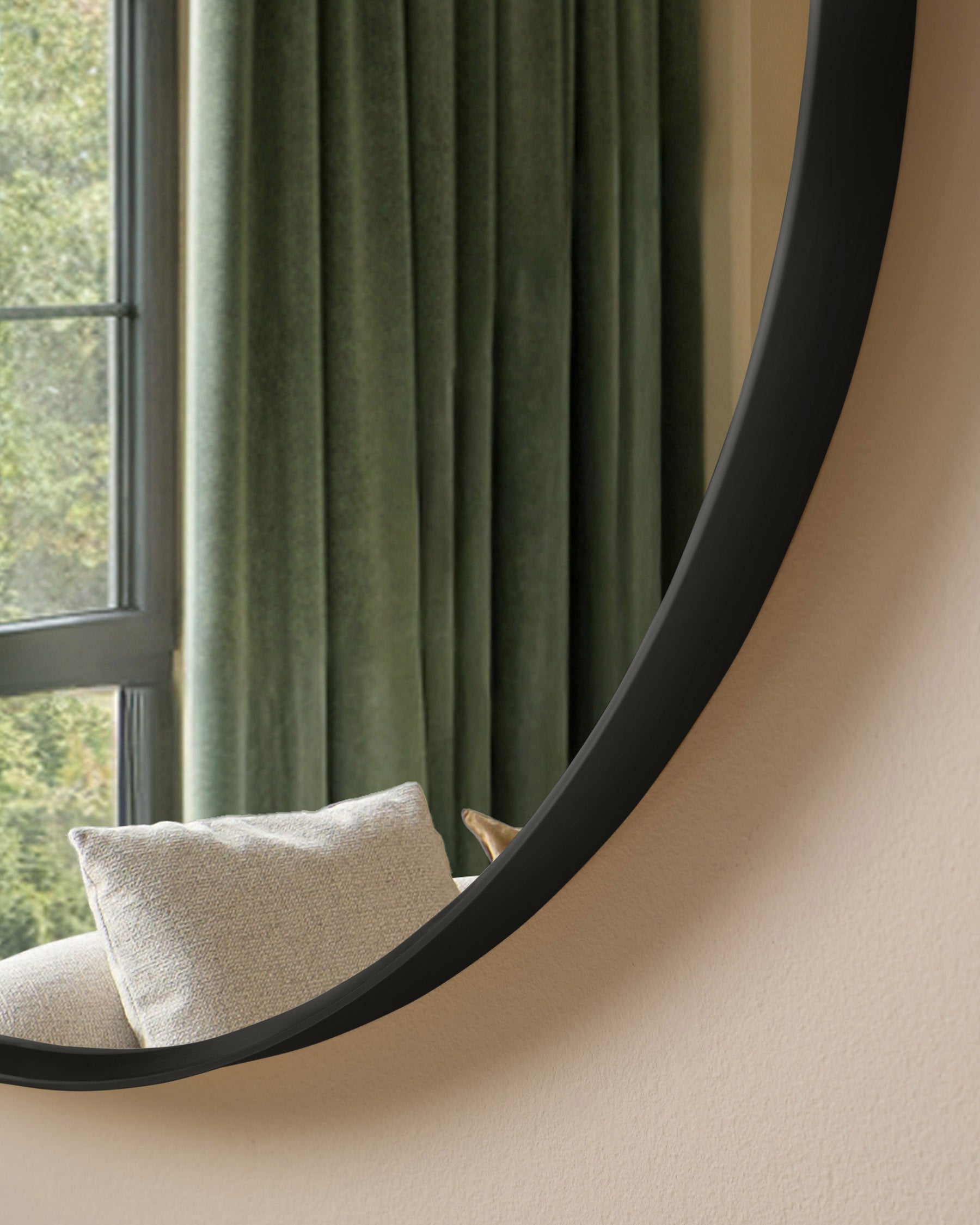 Black curved wall mirror with organic, flowing shape displayed above a cream upholstered chair near a window with green curtains.