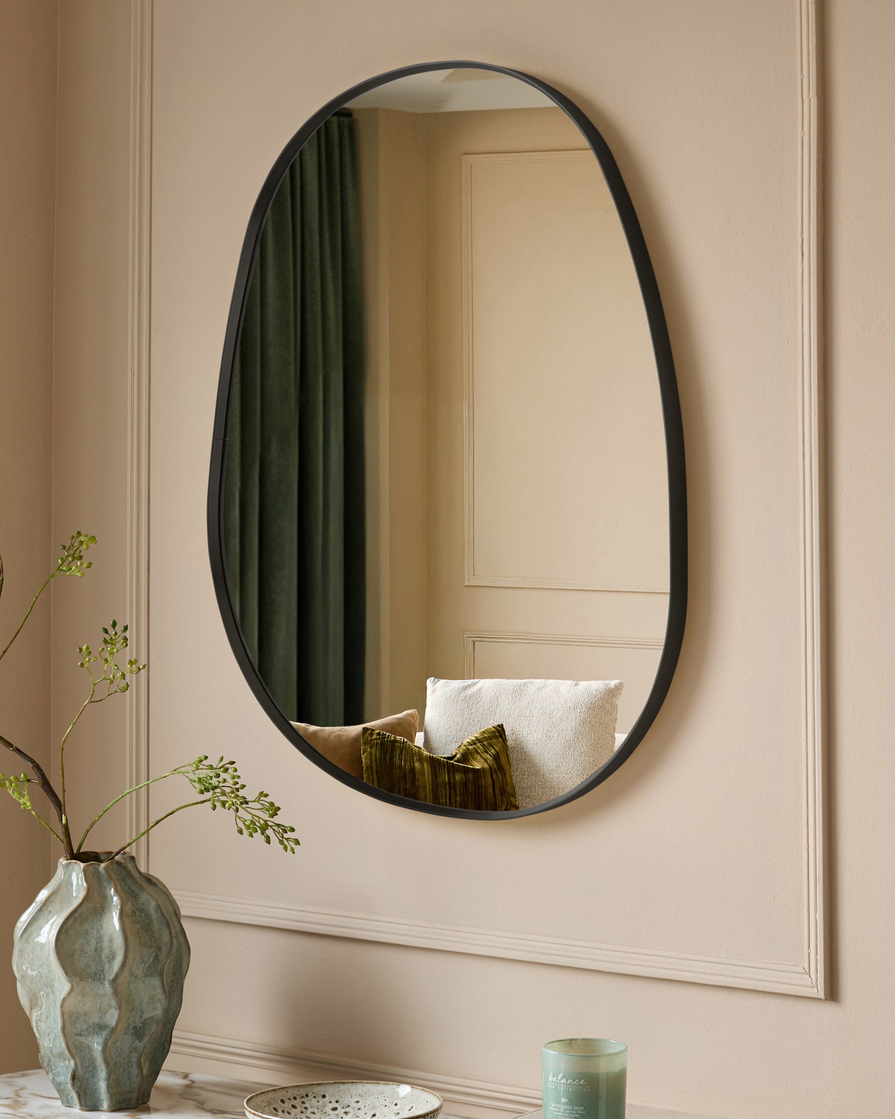 Black oval wall mirror with curved frame, displayed on beige wall above white shelf with decorative vase and accessories.