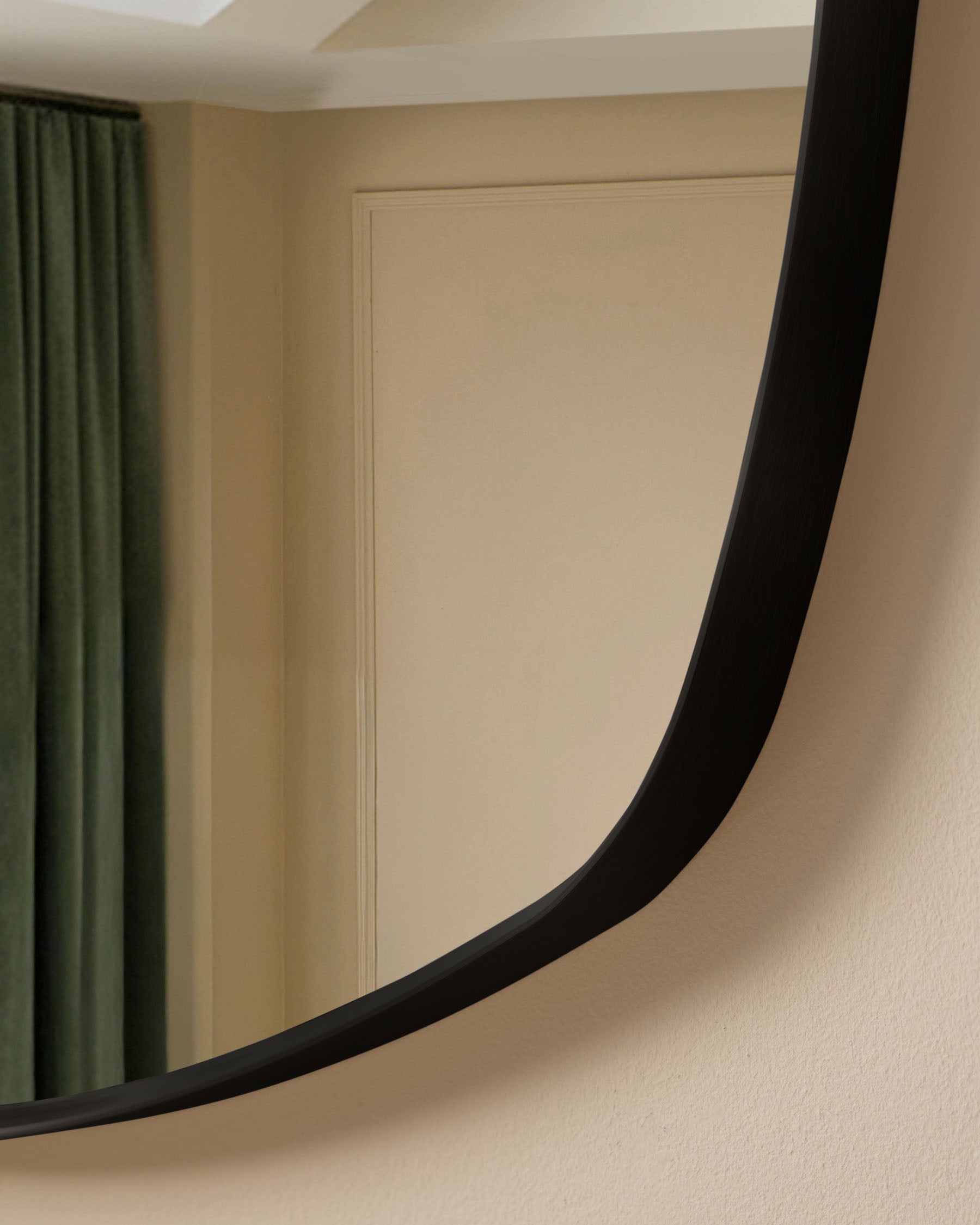 Black curved wall mirror with sleek frame, mounted on beige textured wall with green curtains visible.