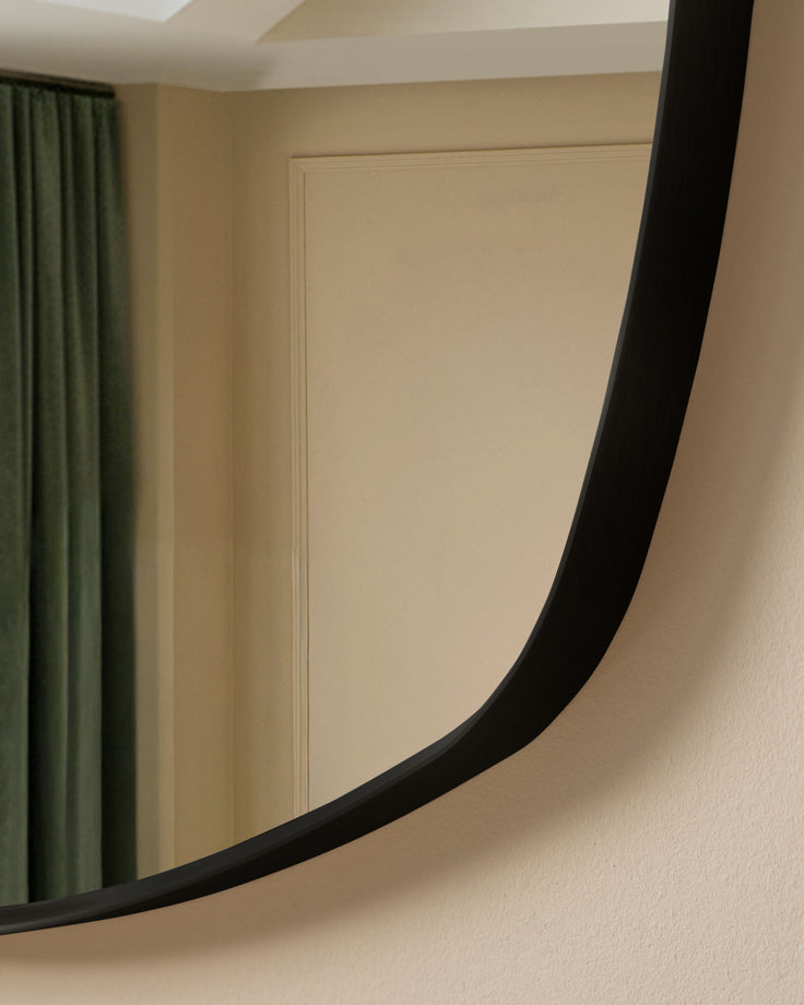 Black curved wall mirror with sleek frame, mounted on beige textured wall with green curtains visible.