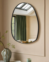 Black oval wall mirror with organic pebble-shaped frame, hung on beige wall above decorative vase with greenery.