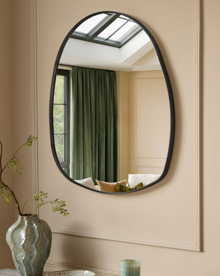 Black oval wall mirror with organic pebble-shaped frame, hung on beige wall above decorative vase with greenery.