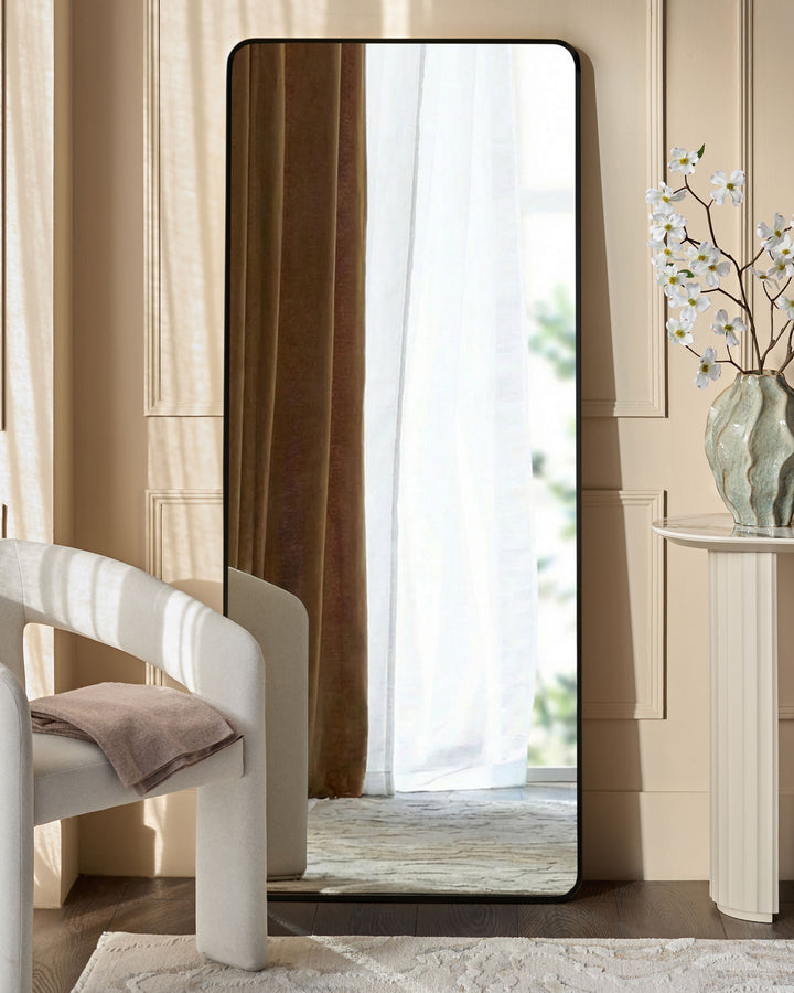 Yannis rectangular full-length leaner mirror with black frame, displayed in a modern bedroom setting against a beige wall.