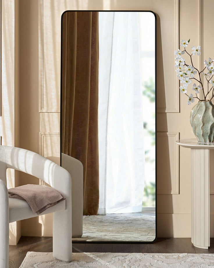 Yannis rectangular full-length leaner mirror with black frame, displayed in a modern bedroom setting against a beige wall.
