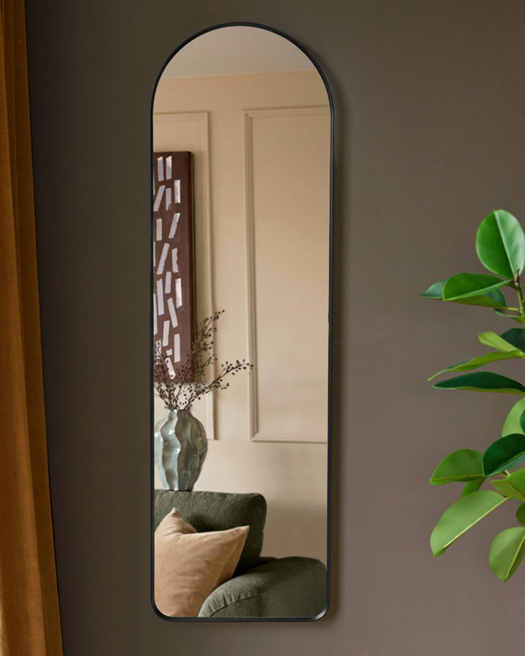 Yannis arch wall mirror with black frame, displaying reflection of modern interior with green chair and decorative vase.