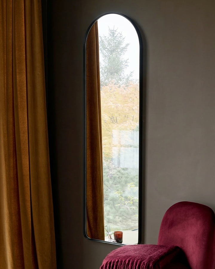 Black arched wall mirror displayed in a modern interior with gold curtains and burgundy chair visible.