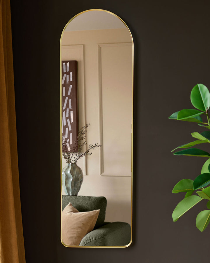 Gold arched wall mirror reflecting interior room with green plant, sofa, and decorative vase on dark wall.