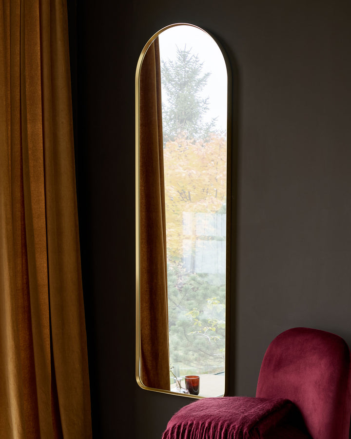 Yannis arch wall mirror with gold finish, tall arched shape, displayed in modern room setting with burgundy chair and gold curtains.