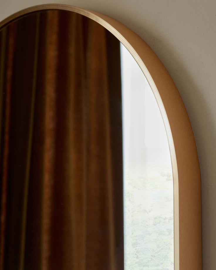 Elegant arched wooden mirror frame with a clear reflection of a window looking out onto green foliage.