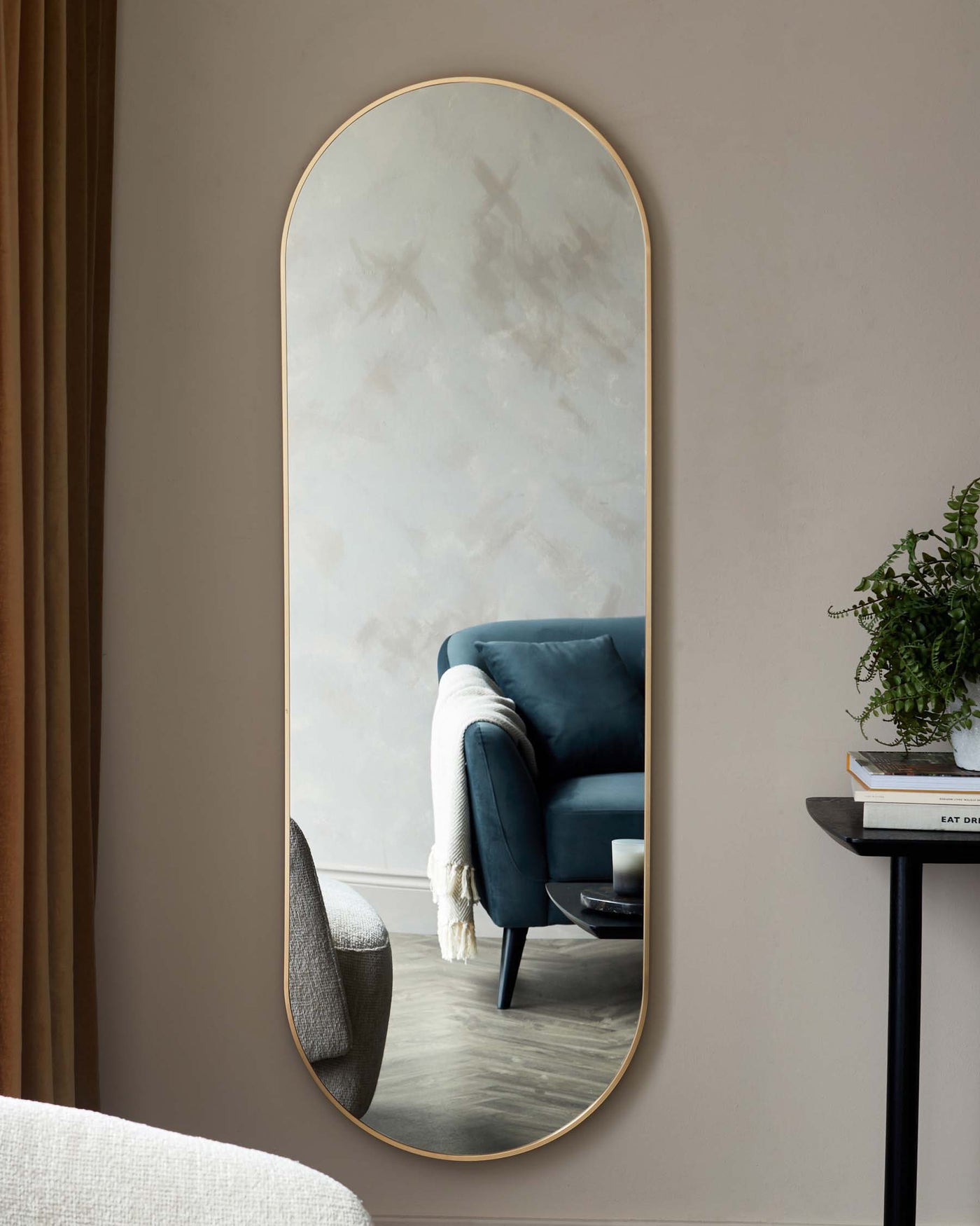 An elegant, tall oval mirror with a slim gold frame is centrally positioned against a soft grey wall. To its right, a modern, circular black side table supports an assortment of books and a lush green potted plant. The reflection captures a luxurious deep blue velvet armchair adorned with a cosy white throw blanket.