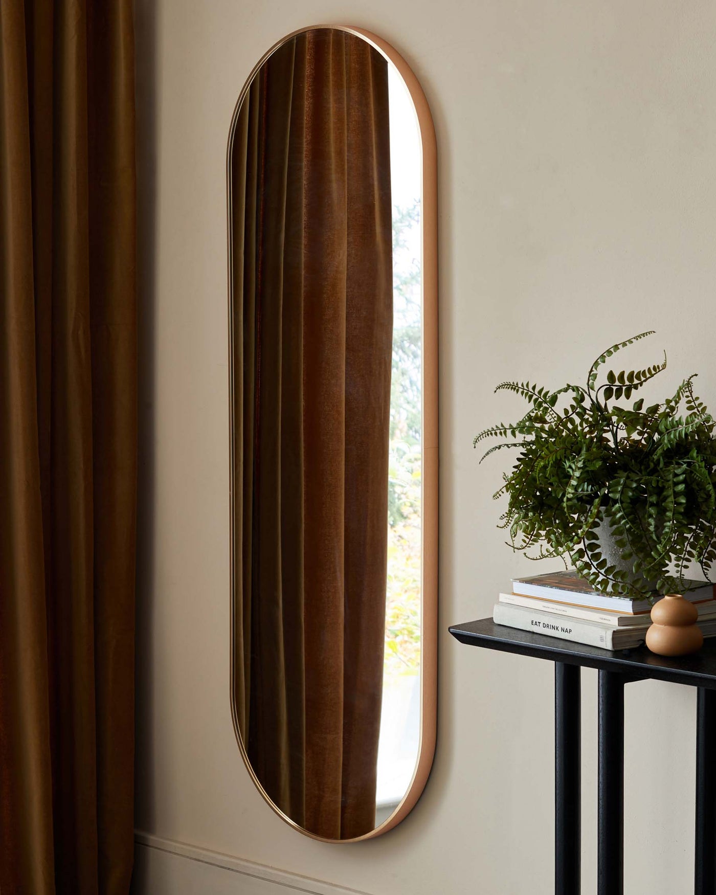 A contemporary, narrow console table with a black metal frame and a smooth, circular top surface, positioned against a light-coloured wall beside an elongated, archtop mirror with a thin, gold-coloured frame. The table is styled with a small stack of books and a decorative spherical object, accompanied by a potted fern to the side.