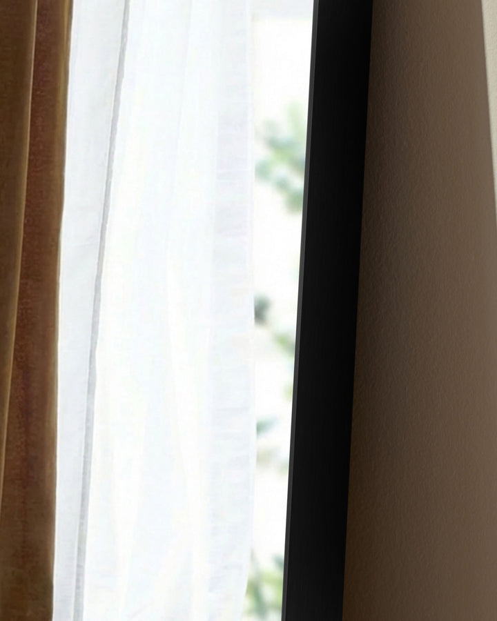 Black rectangular full-length leaner mirror positioned against a wall between curtains, showing its sleek frame and reflective surface.