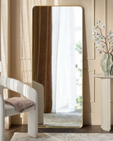Yannis rectangular full-length leaner mirror with gold frame, displayed in a modern bedroom setting against a beige wall.
