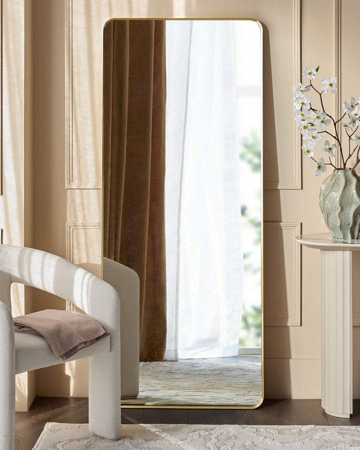 Yannis rectangular full-length leaner mirror with gold frame, displayed in a modern bedroom setting against a beige wall.