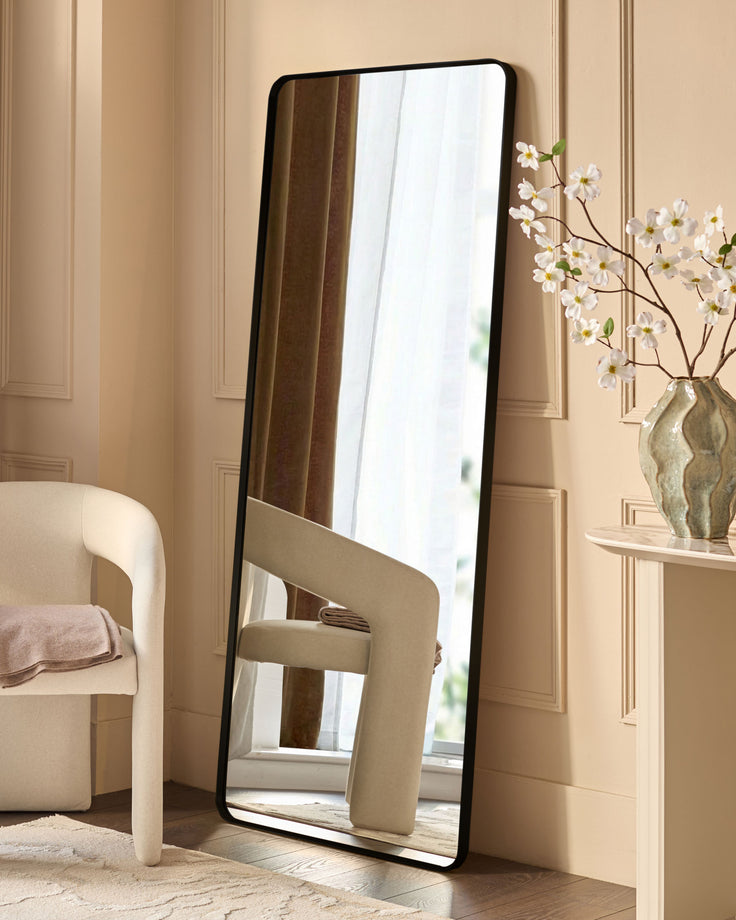 Yannis rectangular full-length leaner mirror with black frame, displayed against a beige wall next to a cream armchair and flowers.
