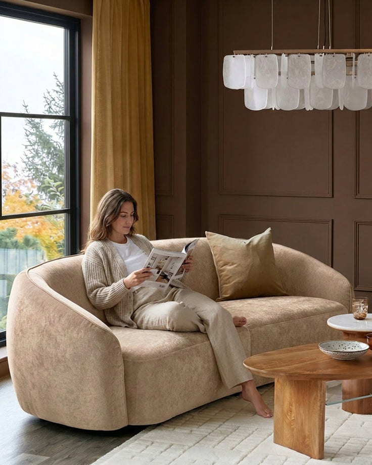 Zoey 3-seater sofa in champagne fabric with curved design, shown in a styled living room setting with a woman reading.