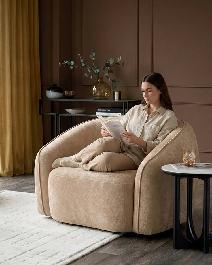 Zoey armchair in champagne fabric, curved design, occupied by woman reading in styled living room setting.
