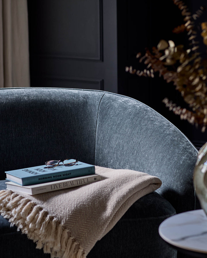 Zoey armchair in blue-grey fabric, curved design, styled with books and throw blanket in modern interior setting.