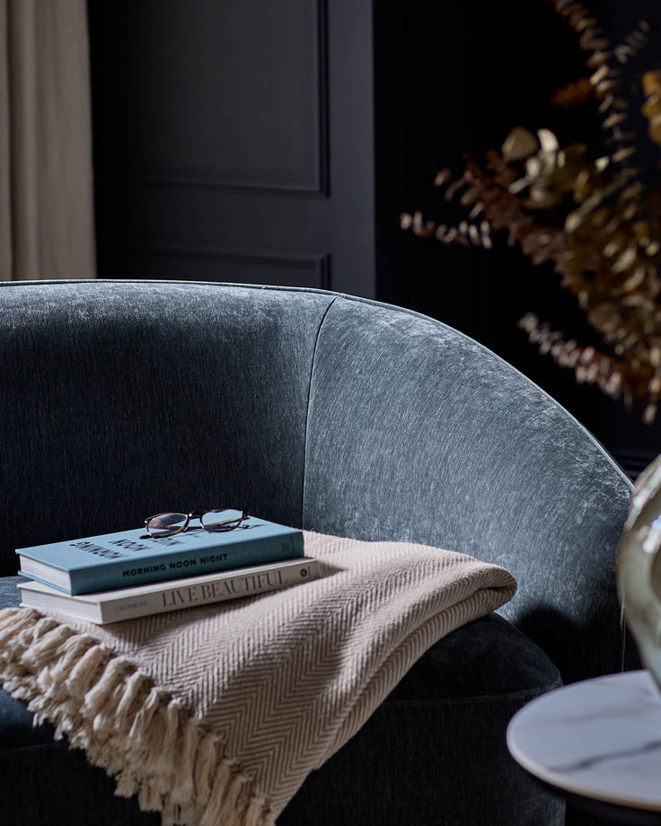 Zoey armchair in blue-grey fabric, curved design, styled with books and throw blanket in modern interior setting.