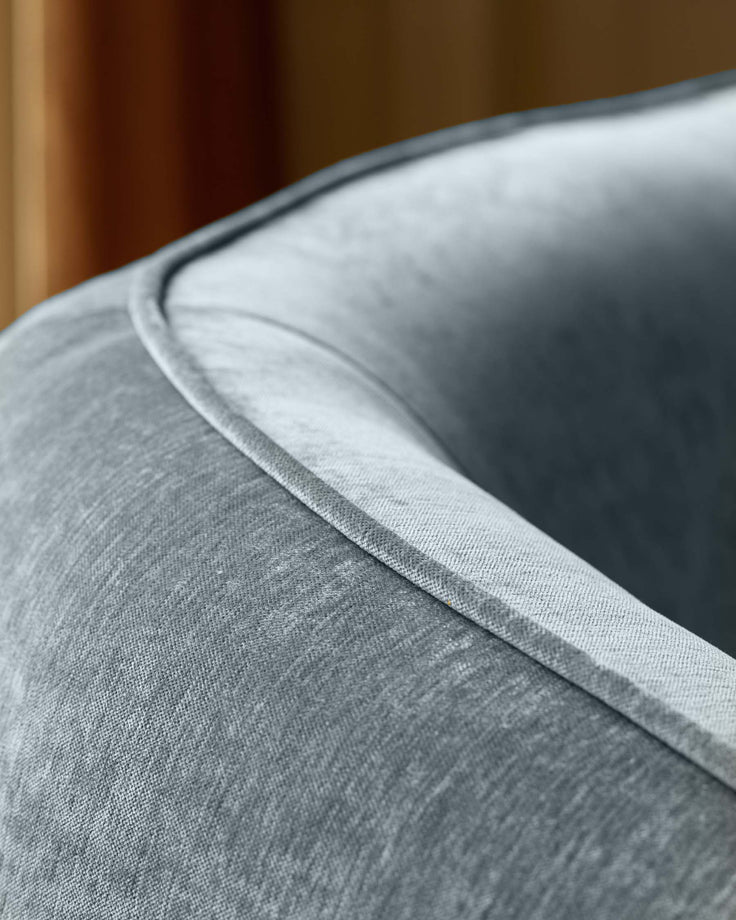 Close-up of a grey armchair, showcasing the fabric texture and piping detail against a blurred background.