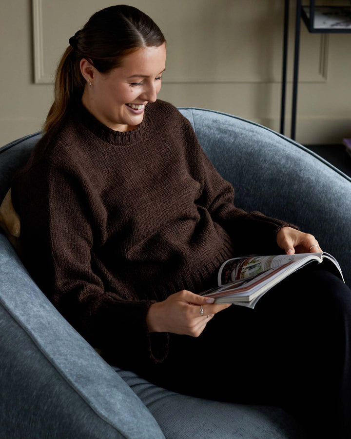 A woman in a cosy brown jumper sits in a blue velvet chair, reading a magazine. A shelving unit is visible in the background.