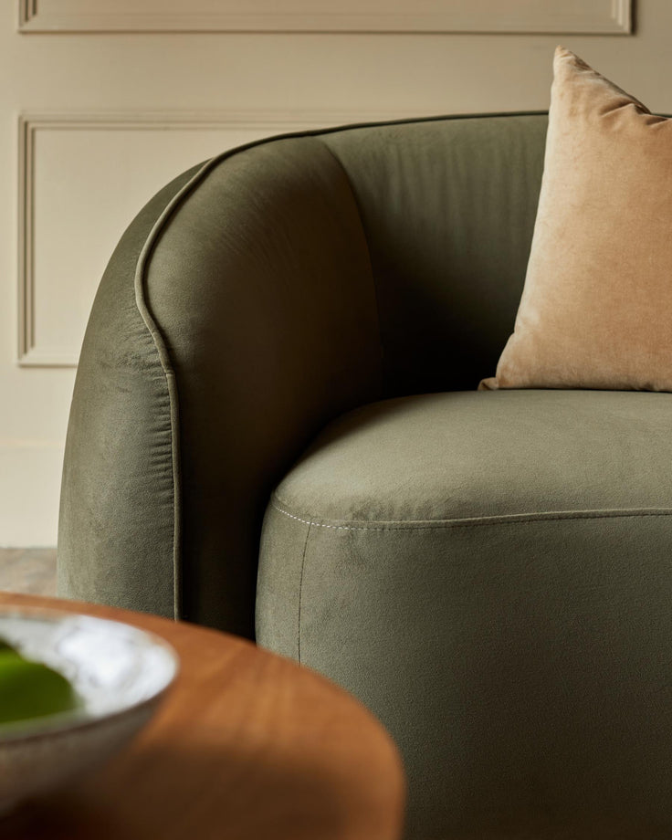 Close-up of an olive green, curved armchair with a beige cushion. Wooden table partly visible in the foreground.