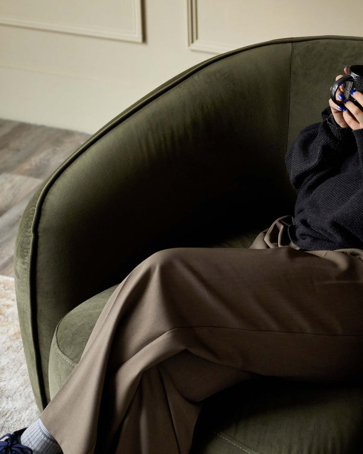 A velvet green armchair with a person sat in it. They're wearing brown trousers and holding a mug.