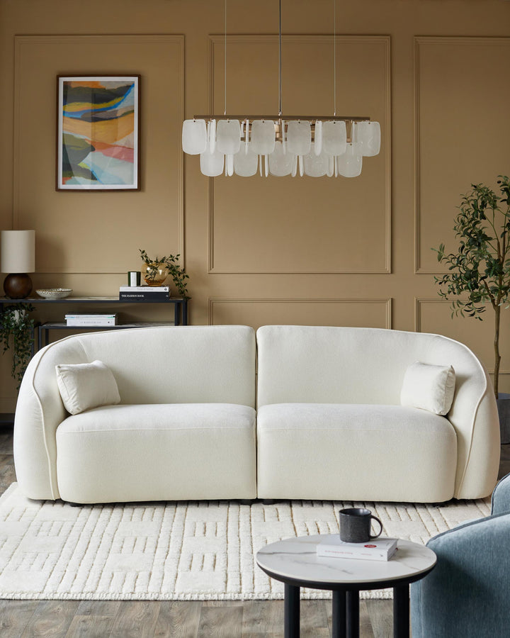 A stylish living room with a cream sofa, patterned rug, modern lighting, and a side table with a mug and book.