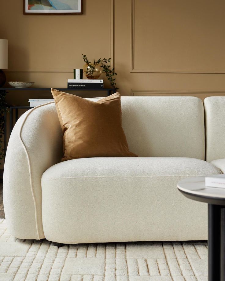 A stylish, cream coloured boucle sofa with a brown velvet cushion sits on a textured rug, next to a black legged table.