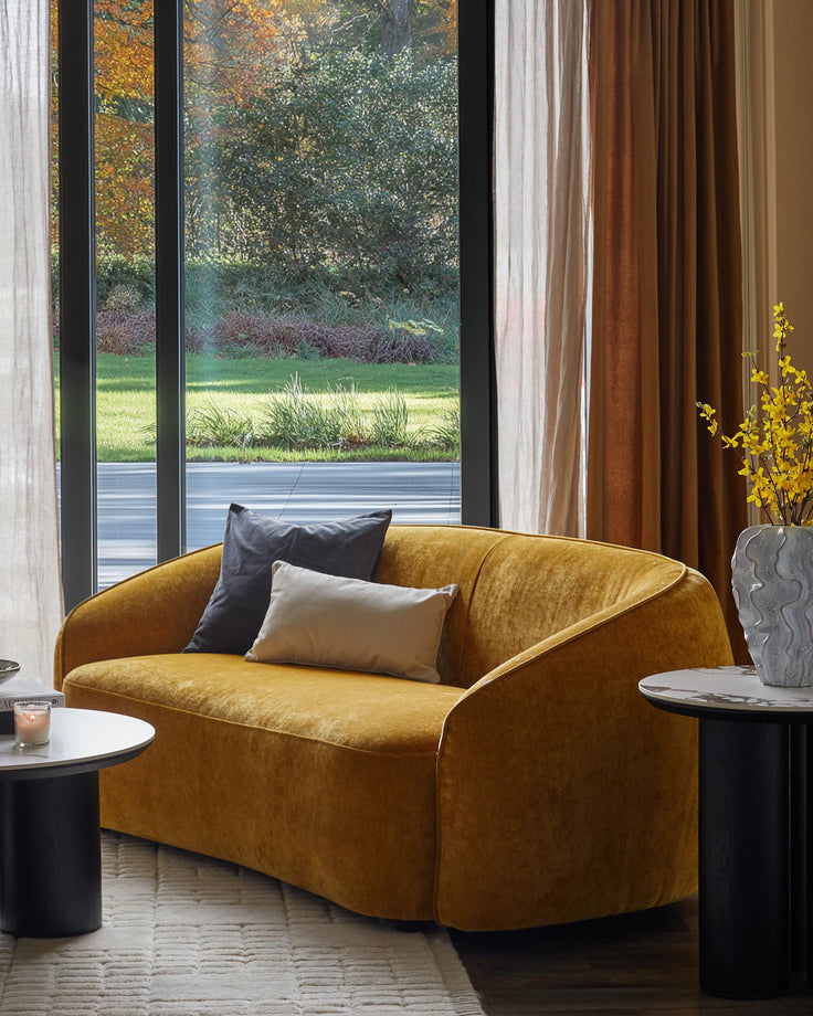 Stylish golden-yellow sofa with cushions, flanked by black and marble side tables. Neutral curtains create a soft backdrop.