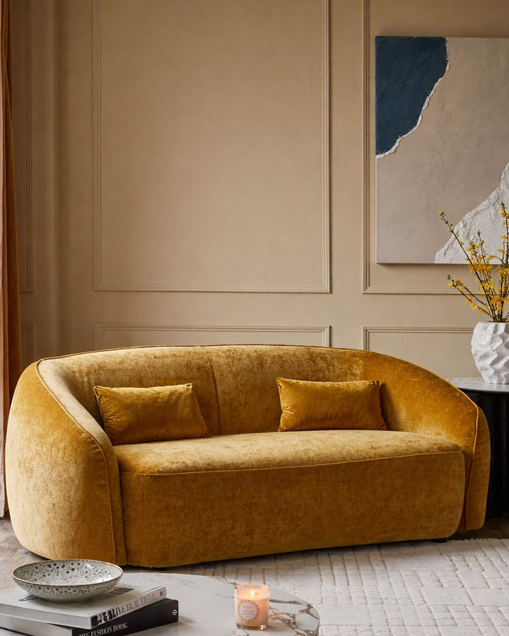 Zoey 2-seater sofa in ochre fabric with curved arms and two matching cushions in a modern living room setting.