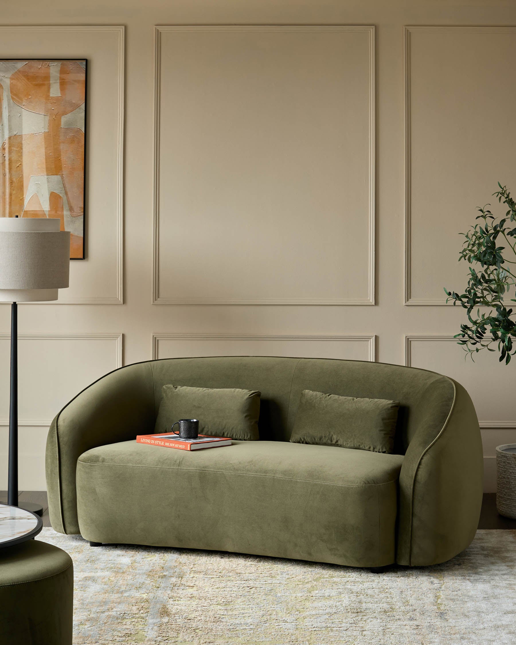 A stylish, olive green velvet sofa with two cushions, alongside a matching stool, on a patterned rug in a neutral room.