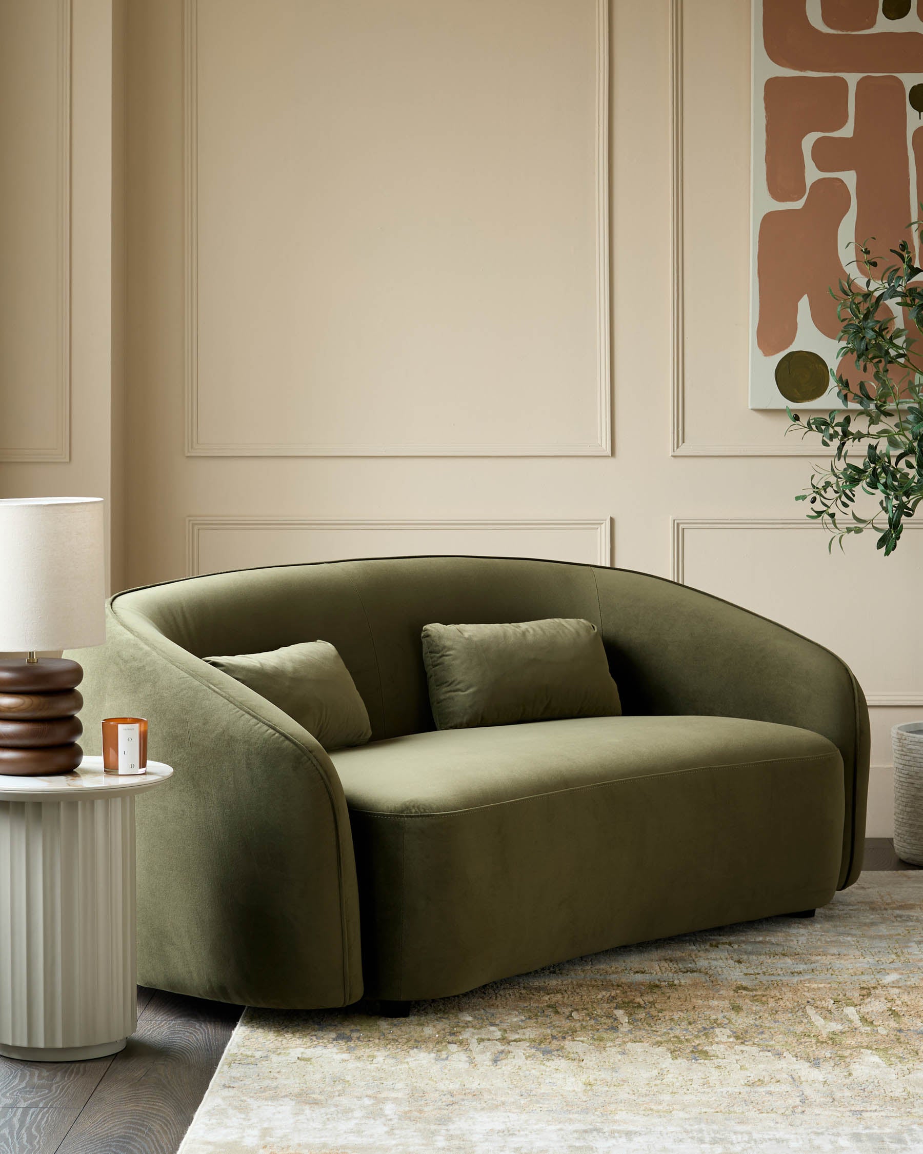 Stylish olive green curved sofa with cushions, side table with lamp, and framed abstract art in a modern living room setting.