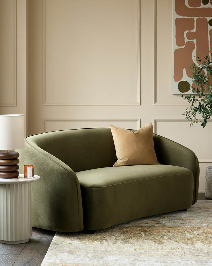 A stylish olive green loveseat with a beige cushion sits next to a ribbed side table and a decorative lamp.