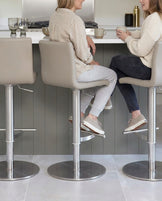Light grey real leather gas lift bar stool with brushed metal base, shown in modern kitchen setting with two people seated.