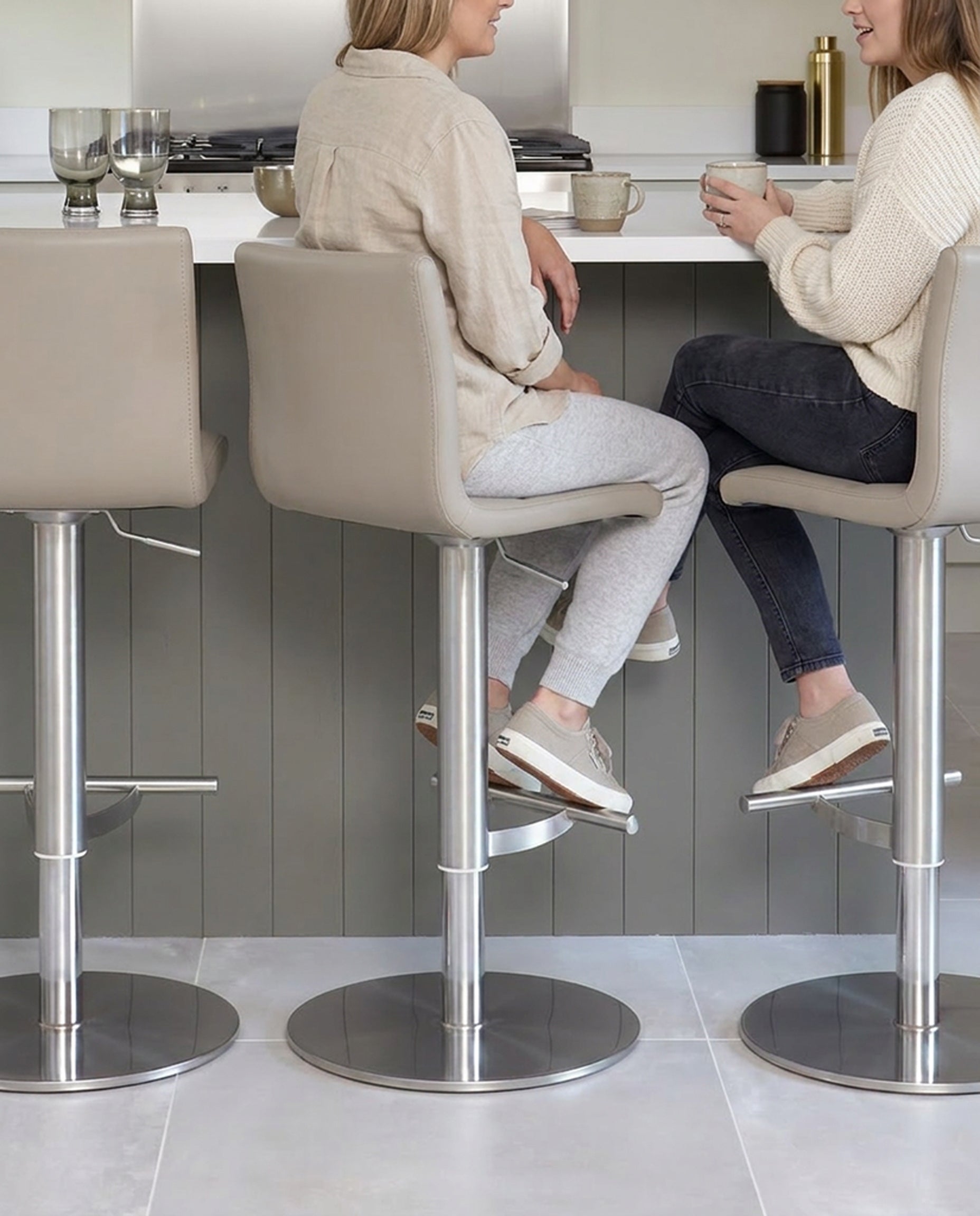 Light grey real leather gas lift bar stool with brushed metal base, shown in modern kitchen setting with two people seated.