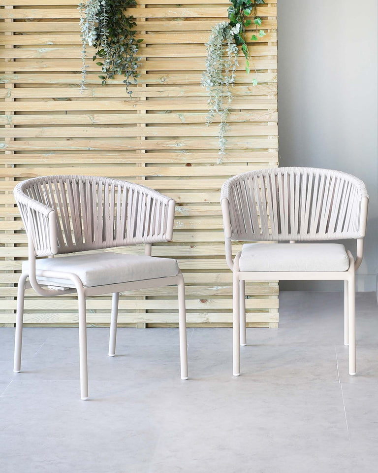 Two contemporary light beige armchairs with a vertical slat back design and upholstered seat pads, set against a wooden slat wall with hanging greenery.