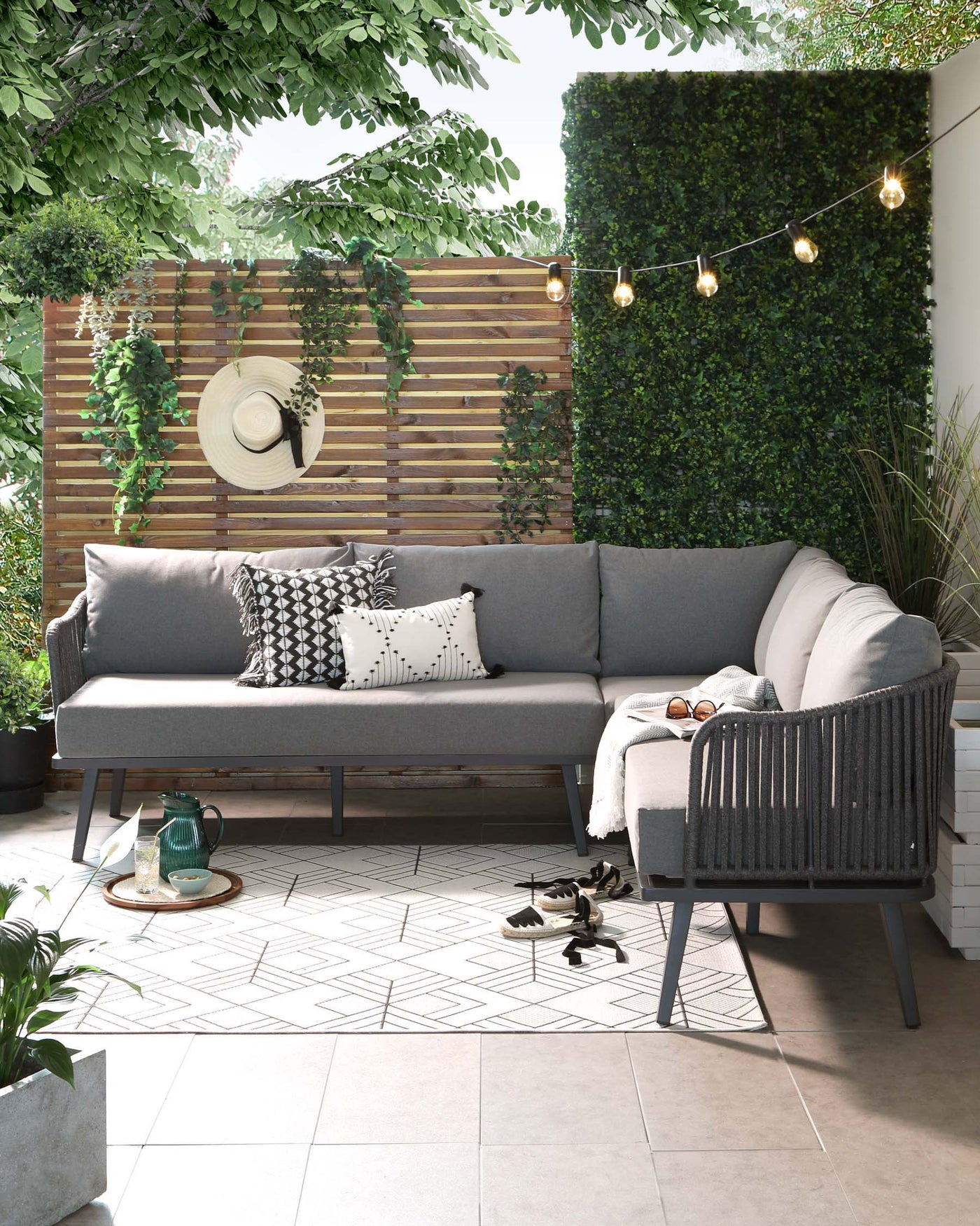 Modern outdoor corner sofa with grey cushions and throw pillows, accompanied by a matching armchair. There is a round wooden side table with a glass pitcher and two cups, set on a geometric-patterned tile floor. Decorative string lights hang above.