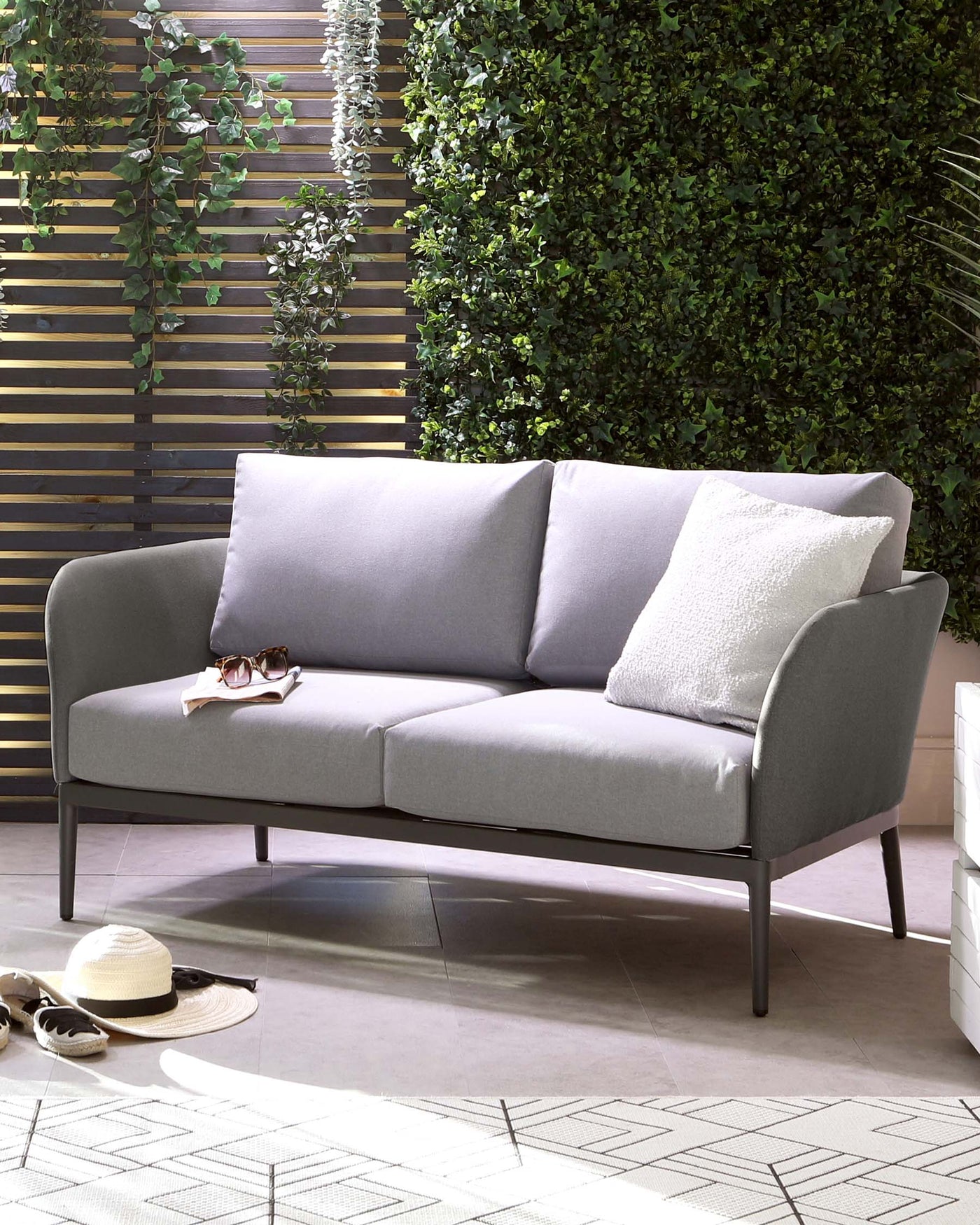 Modern outdoor three-seater sofa with a sleek metal frame and plush grey cushions, accompanied by fluffy white pillows. The piece is accessorized with a pair of sunglasses and a sun hat, evoking a relaxed, chic outdoor living vibe.