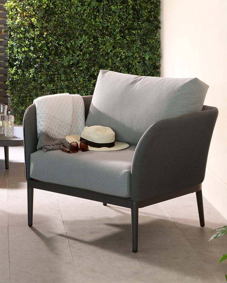 Contemporary two-seater loveseat with grey upholstery and dark slim legs, accented with a patterned light grey throw and a large grey cushion. A straw hat and sunglasses rest on the seat, suggesting a relaxed, stylish vibe. Set against a backdrop of a verdant vertical garden wall.