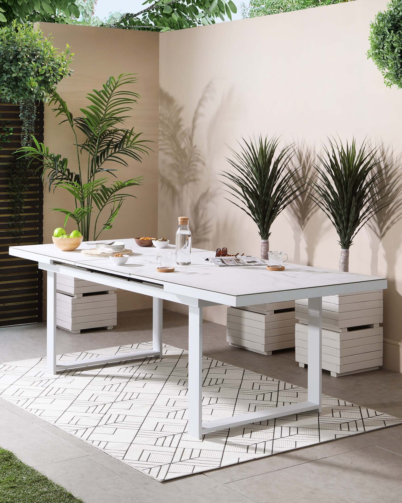 Modern outdoor dining furniture set consisting of a large white rectangular table with a sleek finish and two matching benches without backrests, featuring horizontal slatted design. The set is arranged on a geometric-patterned area rug, enhancing its contemporary appeal. Plants and dining ware are used as accents.