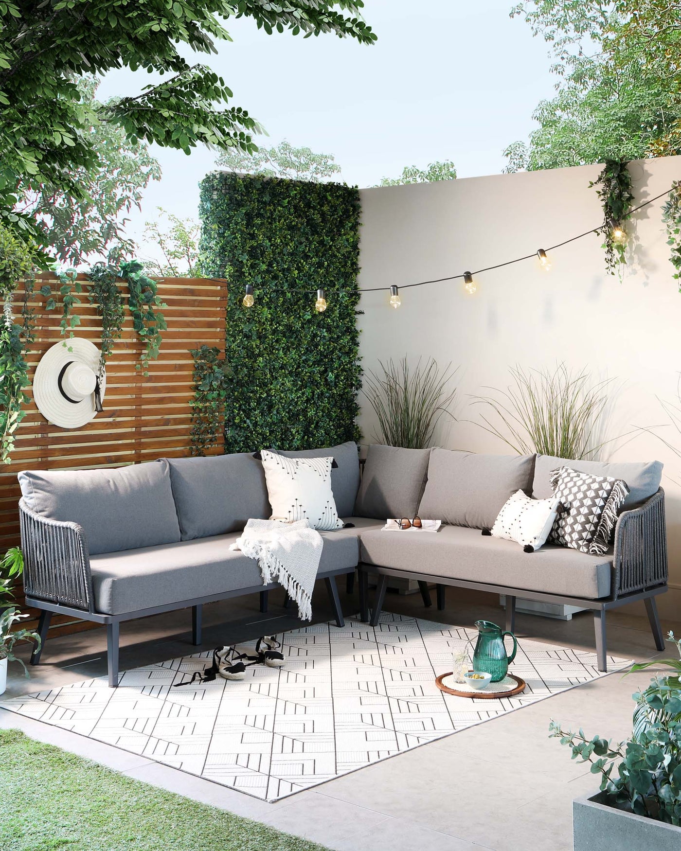 Outdoor corner lounge set with a modern design, featuring a two-piece sectional sofa with comfortable grey cushions, complemented by decorative pillows in various patterns, and a matching low-profile coffee table. The ensemble is arranged on a black and white geometric patterned area rug, enhancing the contemporary aesthetic.