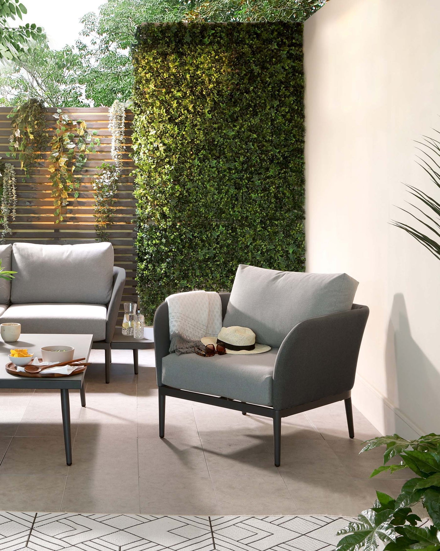 Modern outdoor furniture set with sleek metal frames, including a two-seater sofa and an armchair with clean lines, upholstered in a neutral grey fabric. A matching square low table with a white tabletop is positioned in front of the sofa, holding a tray with a teapot and cups, flanked by assorted decorative items and a small plant.