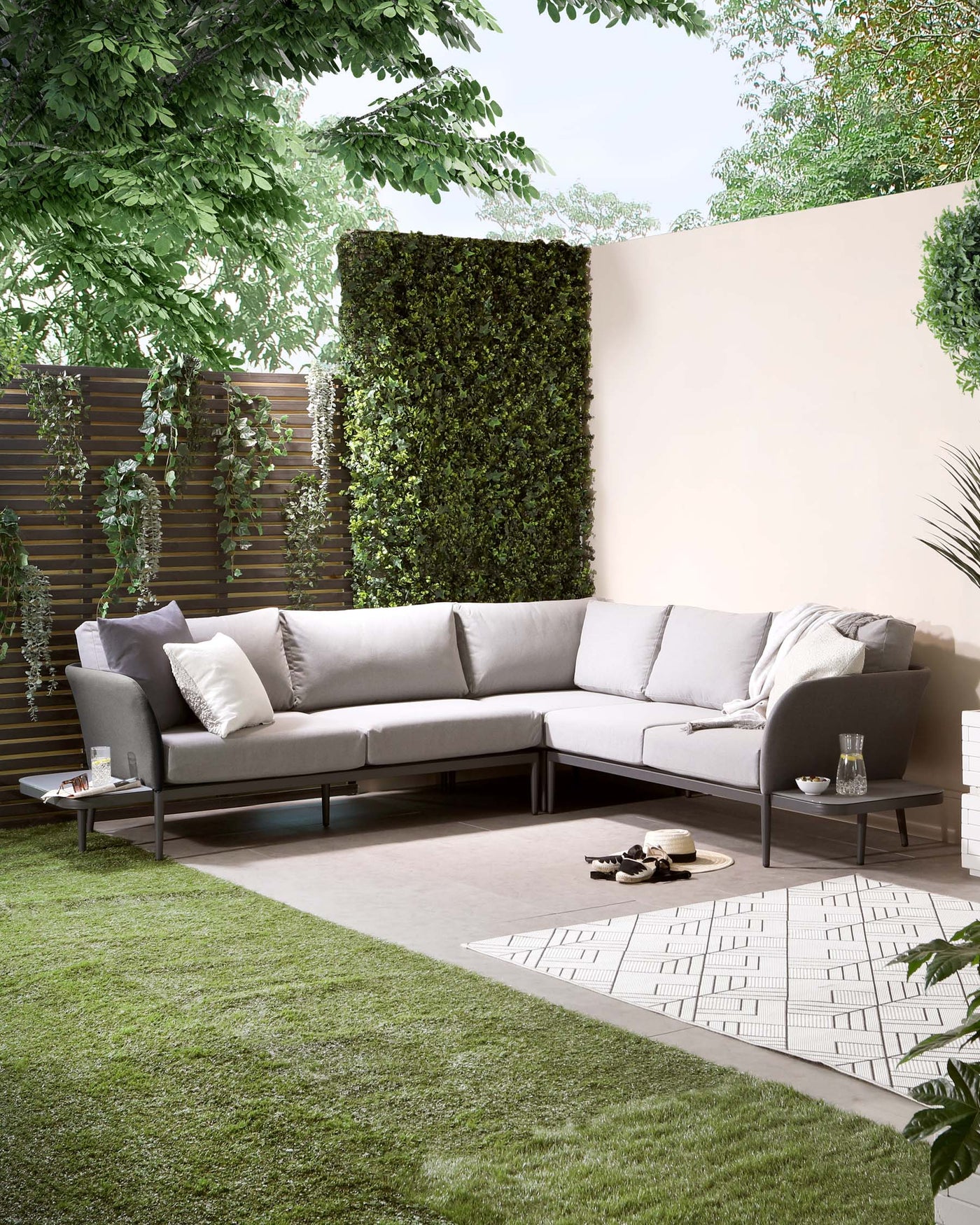 Modern outdoor L-shaped sectional sofa with light grey cushions on a low-profile black metal frame, paired with a small round side table featuring a similar design aesthetic. A decorative grey and white geometric patterned area rug is placed under the sofa.